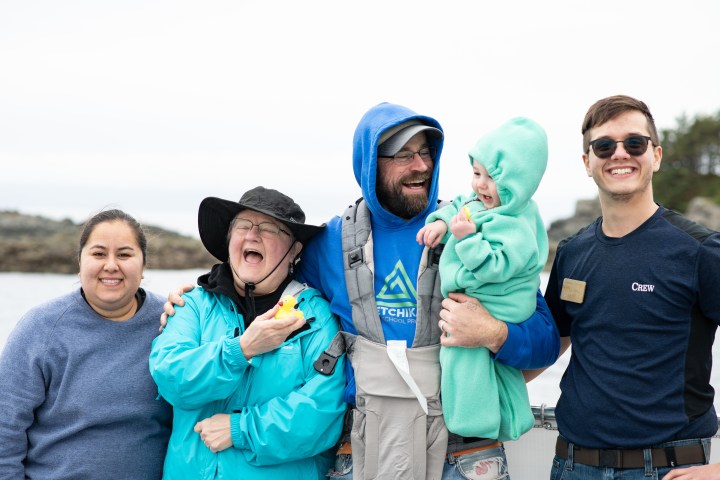Group of five people smiling, one holding a baby and another a rubber duck, with an ocean background.