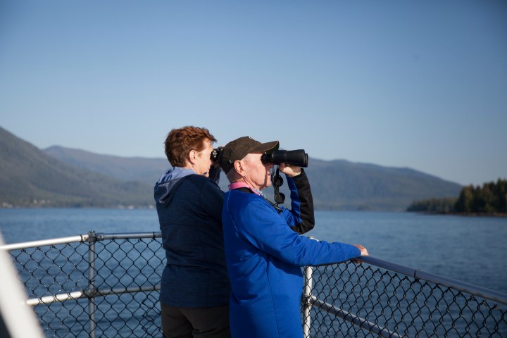 Two people on a boat, one using binoculars, with mountains and water in the background.