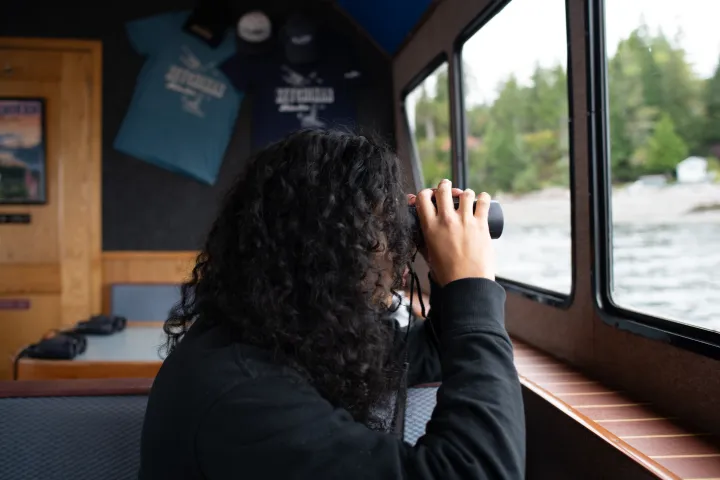 Person with curly hair looking out window using binoculars on a boat.
