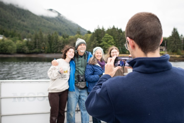 Person photographing four smiling people on a boat with forested mountains in the background.