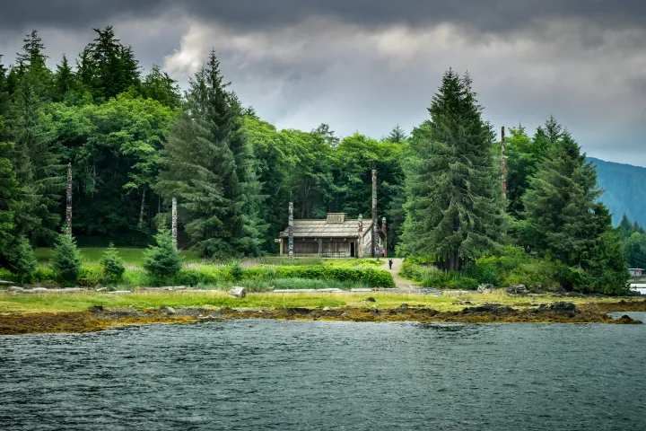Alaska Native clan house and totem poles surrounded by tall trees near a shore under a cloudy sky.
