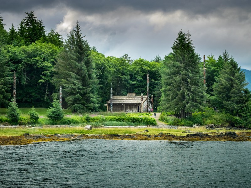 Alaska Native clan house and totem poles surrounded by tall trees near a shore under a cloudy sky.