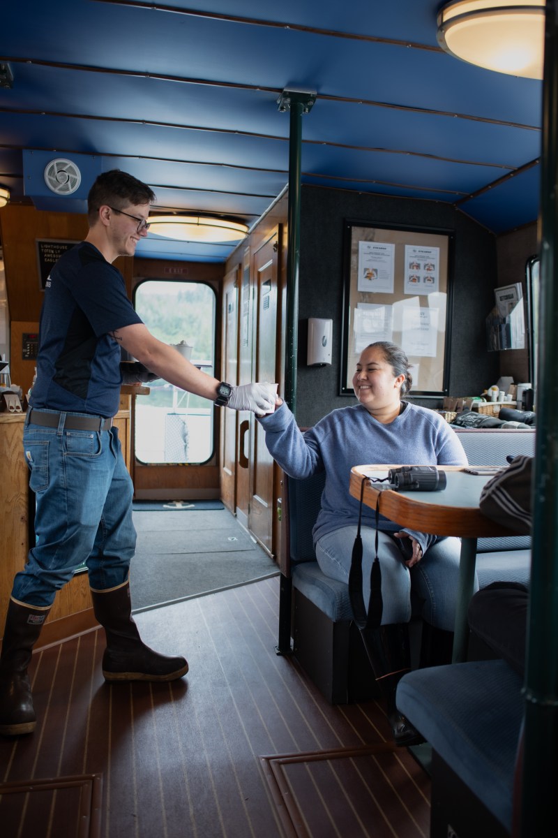 Person in boat cabin handing a cup to a seated woman, both smiling.