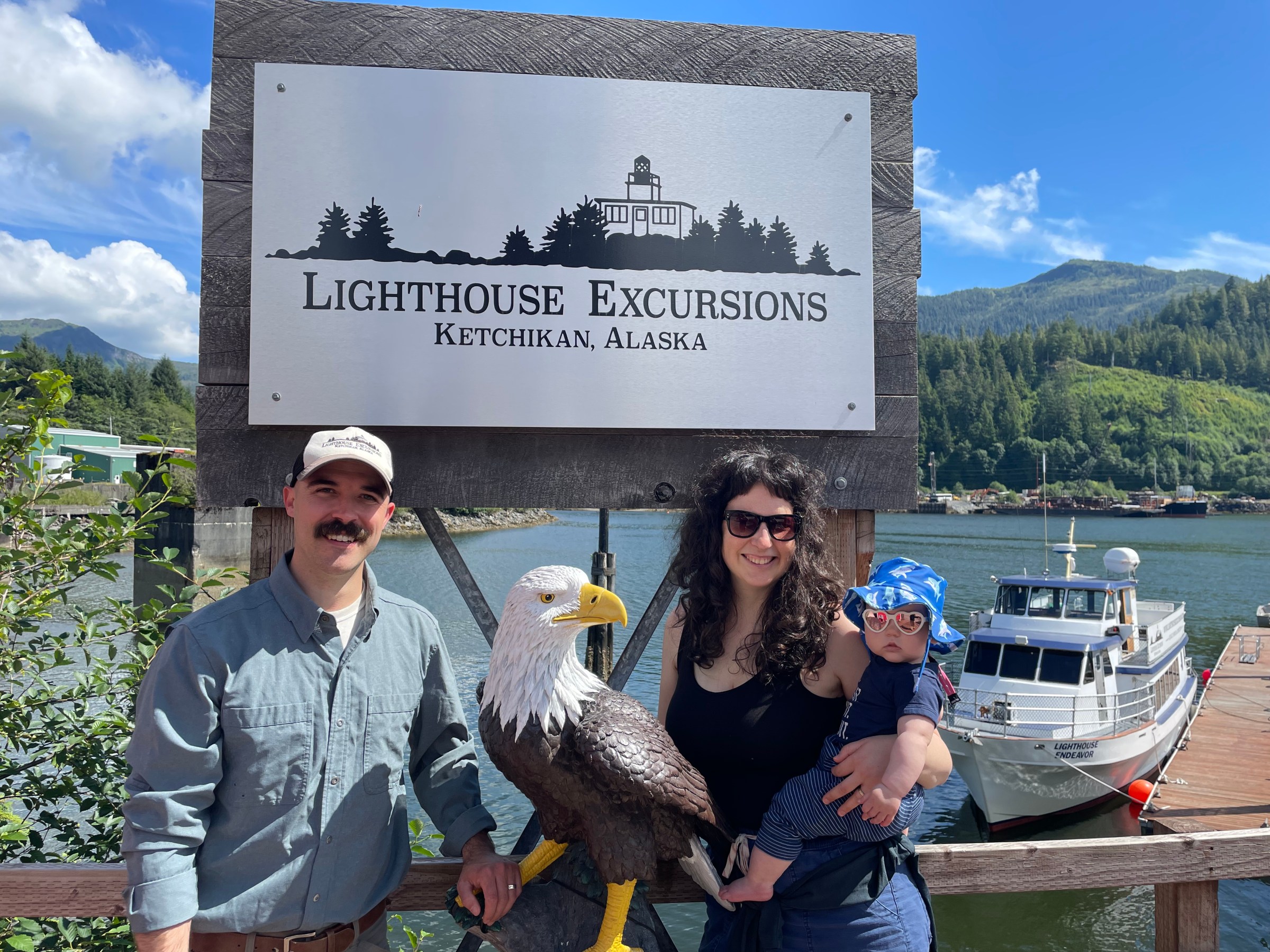 Family poses by 'Lighthouse Excursions' sign with lake and boat in background, Ketchikan, Alaska.