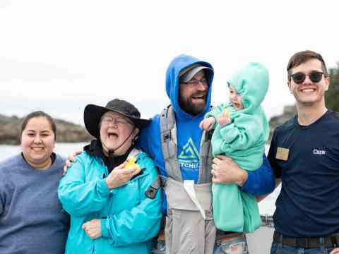 Group of five people smiling on a boat, one holding a child in green suit.