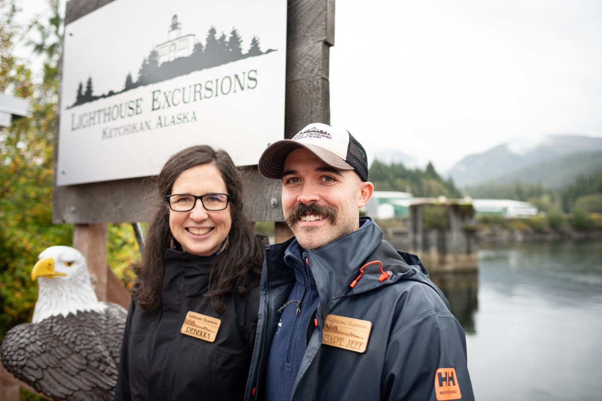 Two people smiling by a sign for Lighthouse Excursions in Ketchikan, Alaska with a bald eagle statue nearby.